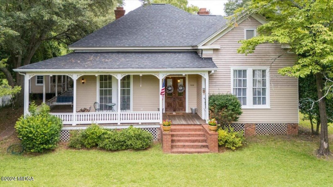 Beige house with a wraparound front porch, brick steps, and an American flag by the front door.