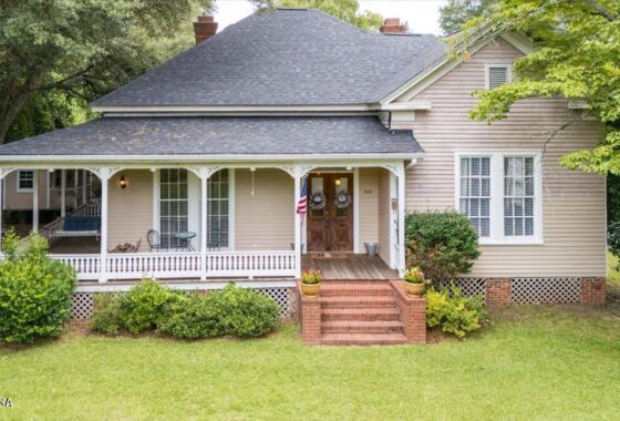 Beige house with a wraparound front porch, brick steps, and an American flag by the front door.