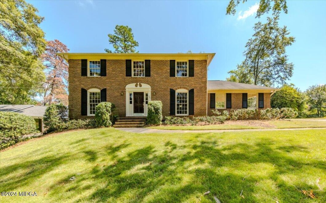 Brick two-story colonial-style house with black shutters, white front door, and manicured shrubs along a curved walkway. A wide lawn and trees surround the property.