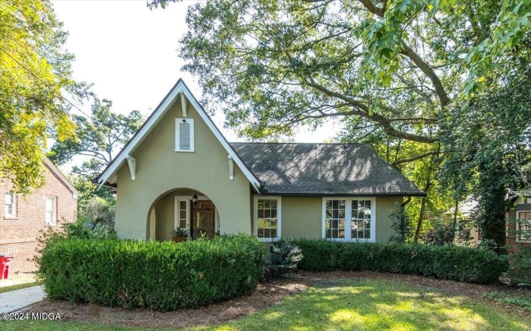 Single-story green stucco house with arched front porch, surrounded by hedges and mature trees.