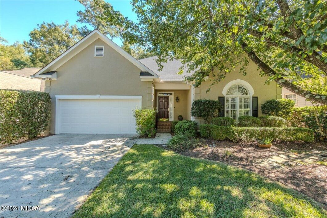 Two-car beige stucco house with white garage door, dark entry door, and manicured shrubs in front yard under a leafy tree