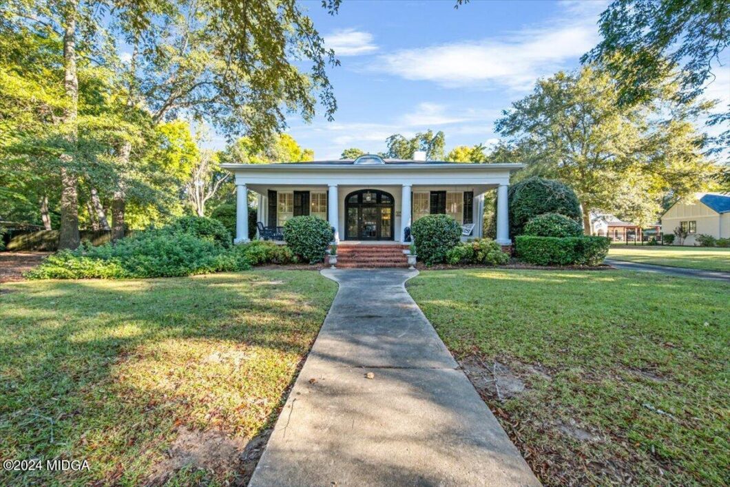 Front view of a white, single-story house with a covered porch supported by four columns and a curved concrete walkway leading from the lawn to the steps.