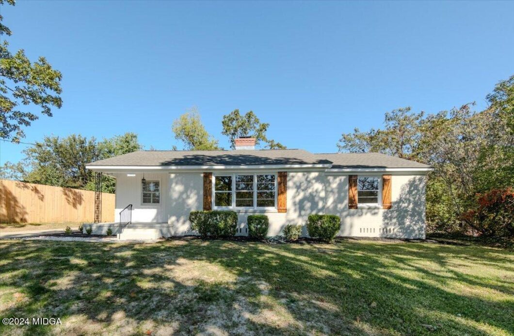 Single-story white ranch-style house with brown window shutters, small front porch, and trimmed shrubs in the yard under a clear blue sky.
