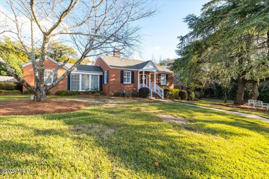 Brick ranch-style house with white trim and a small front porch, surrounded by a well-kept lawn and mature trees under a clear blue sky