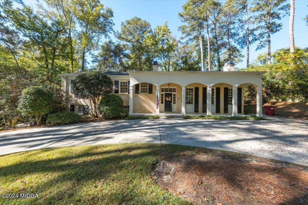 Beige single-story house with a columned front porch, central door with a wreath, an American flag, and a curved driveway bordered by trees nearby.