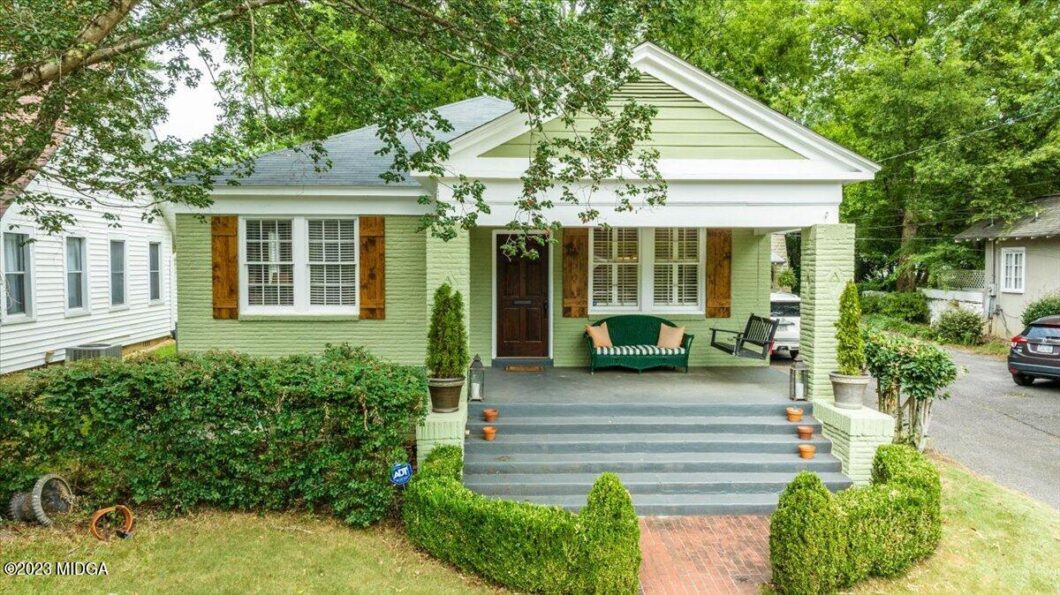Front view of a mint-green cottage with a covered porch, dark front door, and gray steps, surrounded by trees and neatly trimmed hedges.