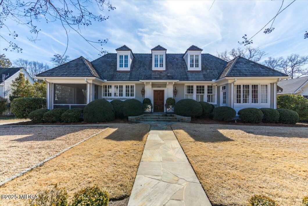 Front view of a white, single-story house with a central wooden door, three dormer windows, and well-manicured rounded shrubs along the walkway.