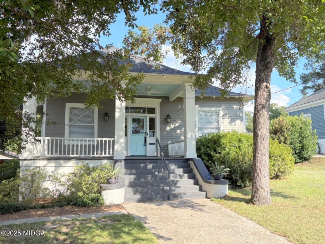 Front porch of a gray house with a light blue door, white columns, and a staircase leading up, shaded by a large tree with greenery around the yard.