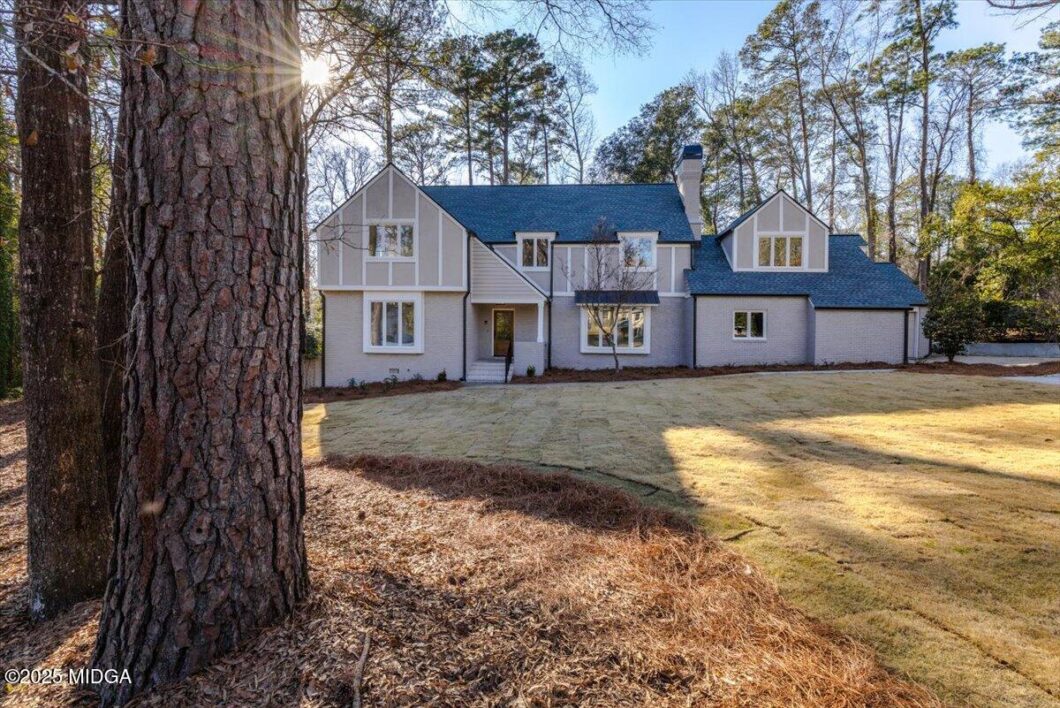 Large two-story house with gray brick and white trim, blue roof, set in a wooded yard with tall trees and a sunlit lawn behind.