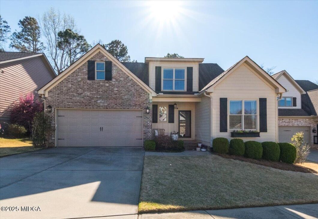 Front view of a two-story suburban house with brick and beige siding, a double garage, and trimmed bushes under bright sun.