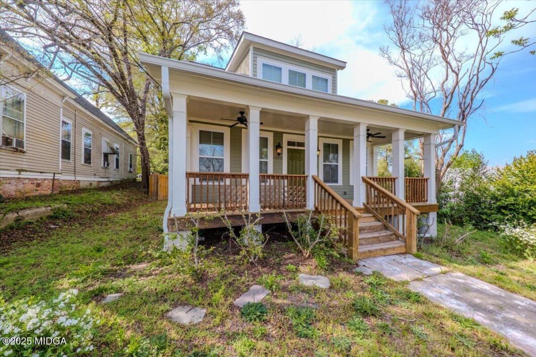 Front view of a light-colored house with a spacious front porch, white columns, wooden railings, and steps up to the door.