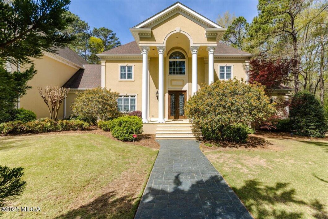 Two-story beige house with tall white columns at the entrance and a curved pediment, surrounded by shrubs and a stone walkway to the front steps.