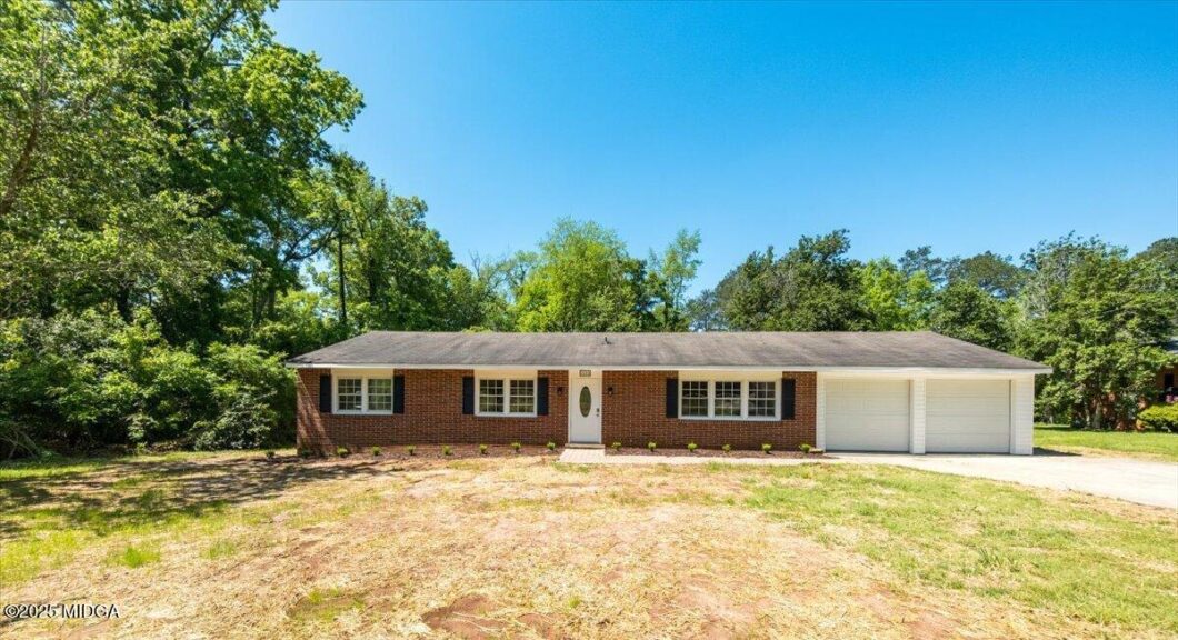 Front view of a single-story brick house with a two-car garage, white door with oval window, and small shrubs along the foundation under a bright blue sky.