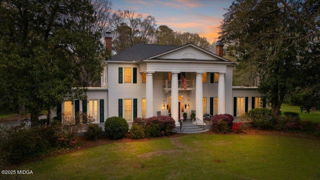 White two-story colonial house with a columned front porch, illuminated windows, and surrounding trees at dusk.