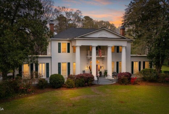 White two-story colonial house with a columned front porch, illuminated windows, and surrounding trees at dusk.