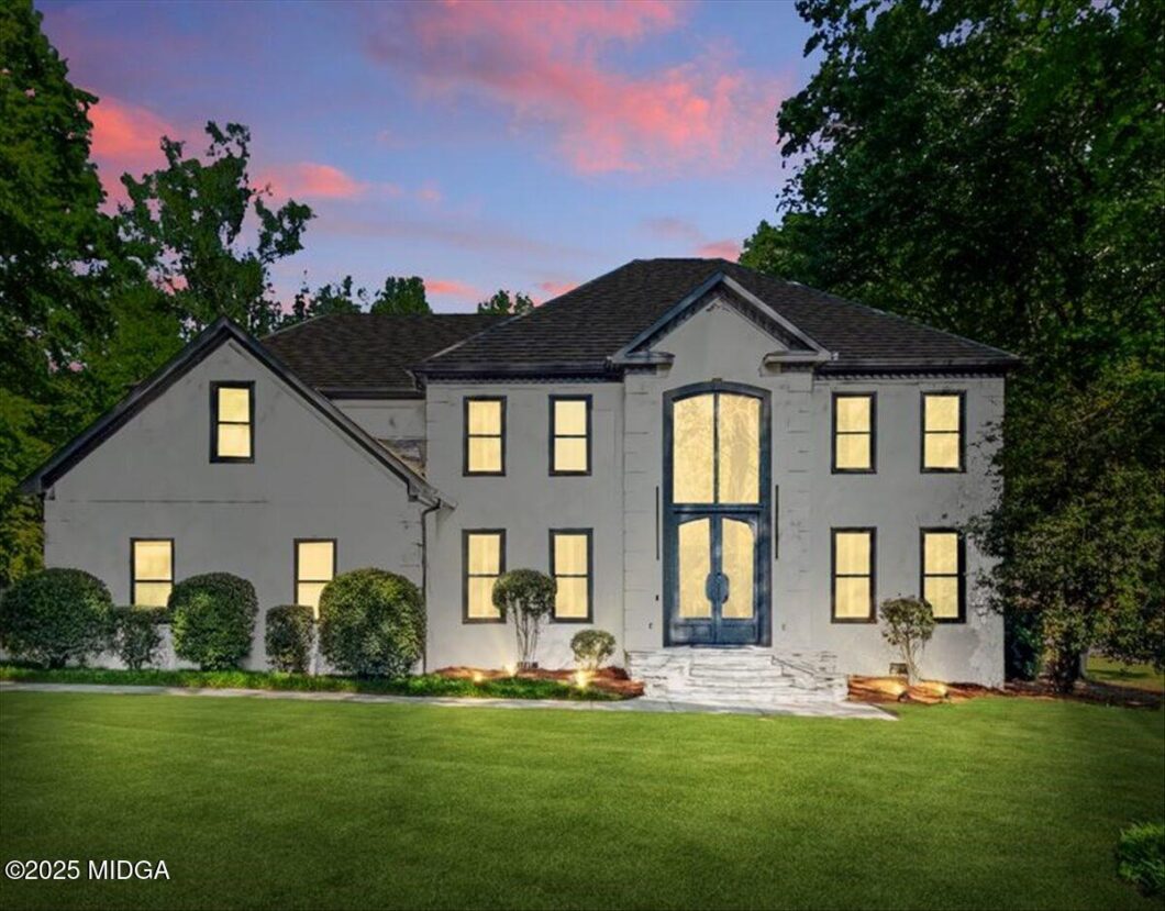 Front view of a white two-story house with illuminated windows, a grand central door, and trimmed shrubs on a lush lawn at dusk.