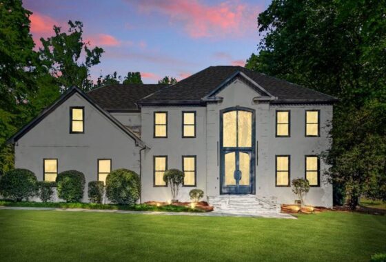 Front view of a white two-story house with illuminated windows, a grand central door, and trimmed shrubs on a lush lawn at dusk.