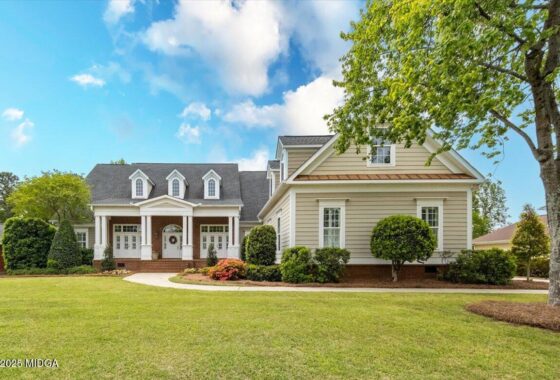 Two-story beige suburban house with a brick-front porch, three dormer windows, and a curved walkway with shrubs. (front view)