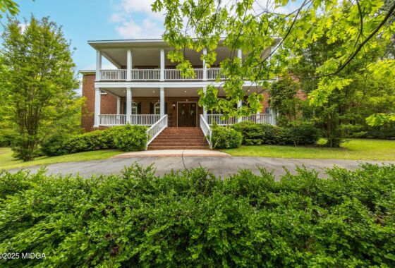 Brick two-story home with wide front porch, white railings, and brick steps surrounded by green shrubs and trees.