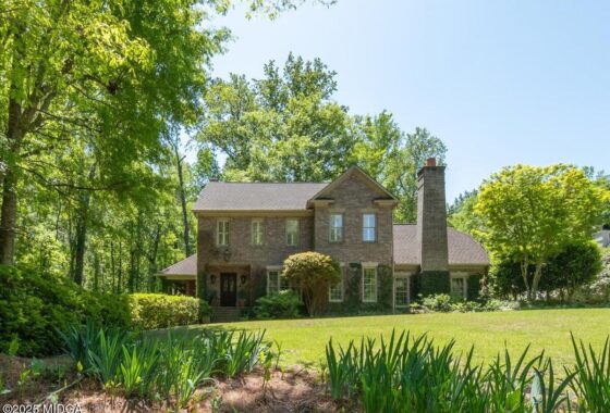 Brick two-story house with a tall chimney, set in a sunny yard with mature trees and a well-kept lawn.