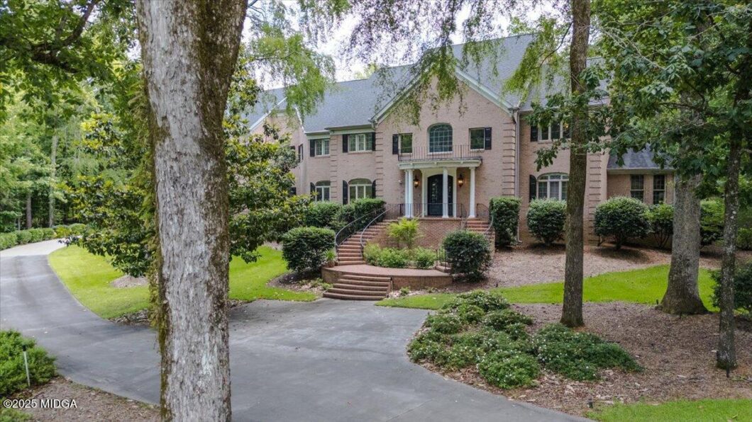 Brick, two-story house with arched windows and a curved brick staircase leading to the front door, set in a wooded landscape with shrubs and trees.