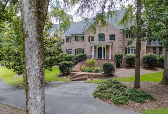 Brick, two-story house with arched windows and a curved brick staircase leading to the front door, set in a wooded landscape with shrubs and trees.
