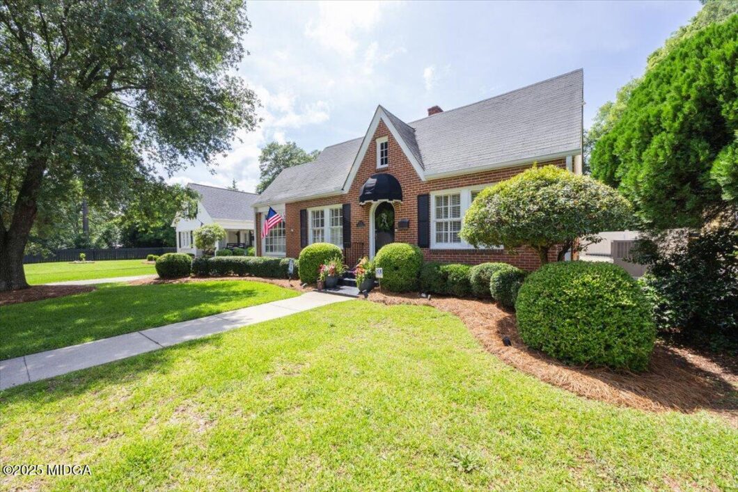 Brick cottage-style house with manicured shrubs, American flag, and a stone path leading to the front door. (Front view of a suburban home)