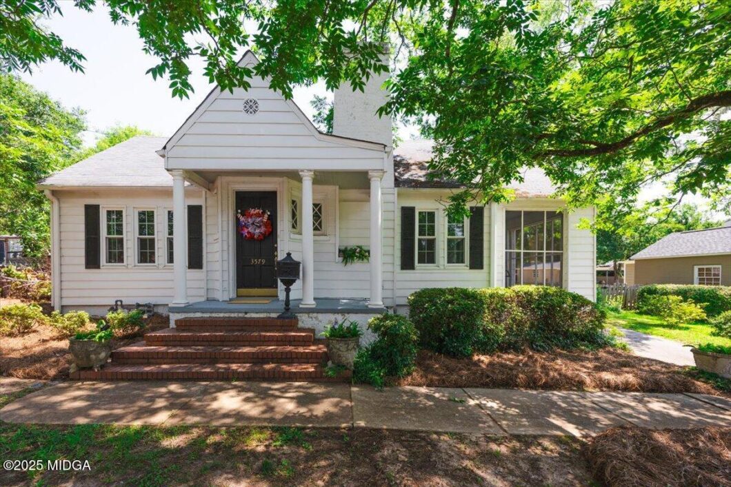 White one-story house with front porch, brick steps, black door with a red wreath, and surrounding green shrubs under a leafy tree