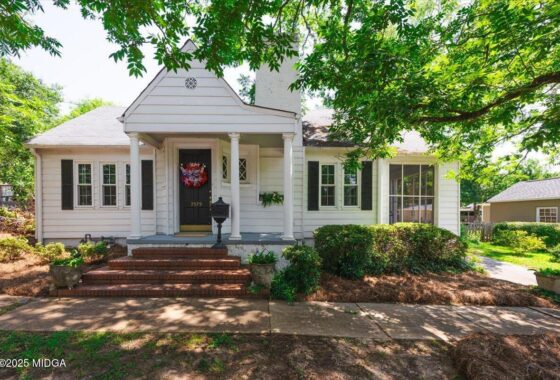 White one-story house with front porch, brick steps, black door with a red wreath, and surrounding green shrubs under a leafy tree