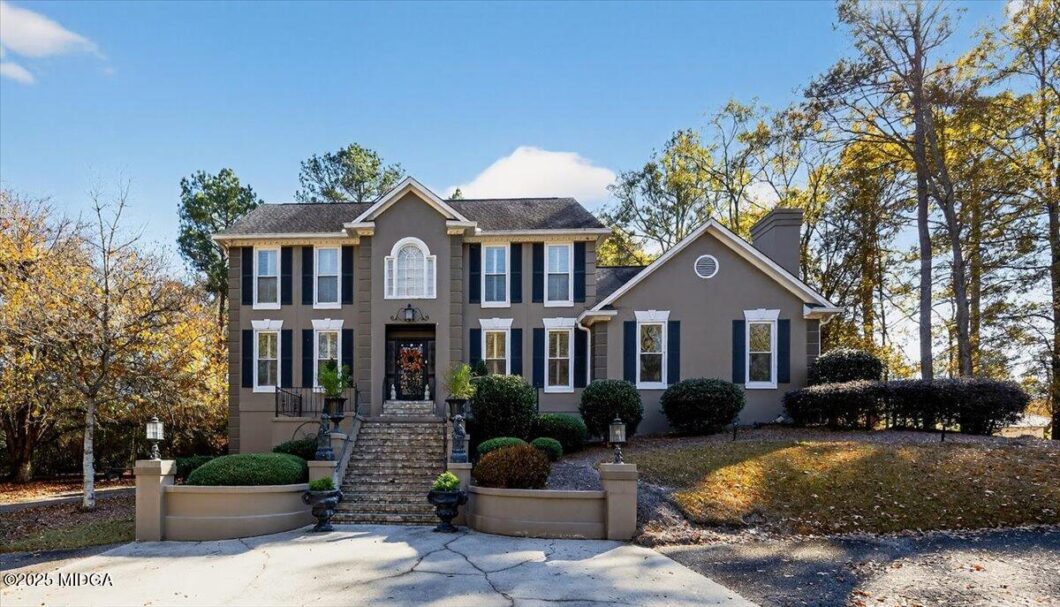Front view of a two-story gray house with black shutters, a central arched window, and stone steps leading to the entrance, flanked by manicured shrubs on a sunny autumn day.