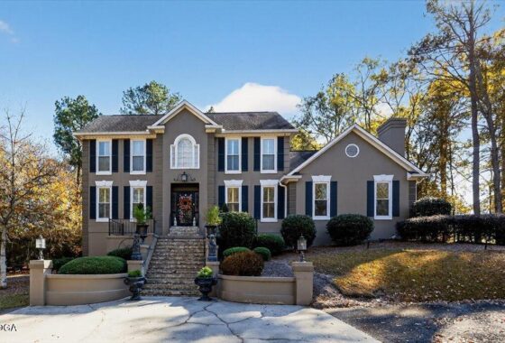 Front view of a two-story gray house with black shutters, a central arched window, and stone steps leading to the entrance, flanked by manicured shrubs on a sunny autumn day.