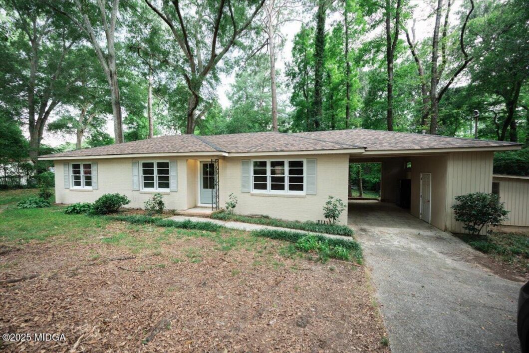 Single-story beige house with a small front porch and attached carport, set among tall green trees in a wooded yard.