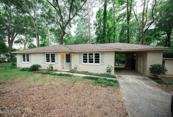 Single-story beige house with a small front porch and attached carport, set among tall green trees in a wooded yard.