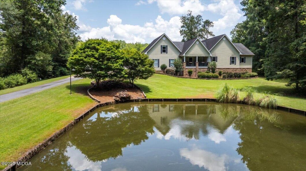 Suburban house with gray siding and brick foundation beside a pond, surrounded by green lawn and trees on a sunny day.