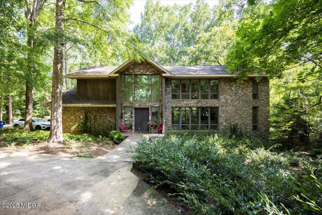 Two-story stone house in a wooded yard with large front windows and a dark front door, surrounded by greenery.