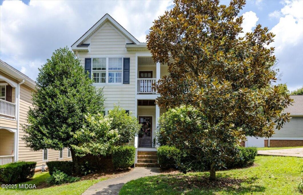 Two-story beige house with white trim and a small front porch, framed by large trees and shrubs in a suburban yard.