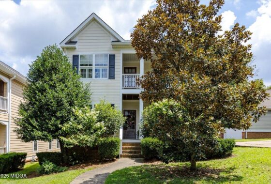 Two-story beige house with white trim and a small front porch, framed by large trees and shrubs in a suburban yard.