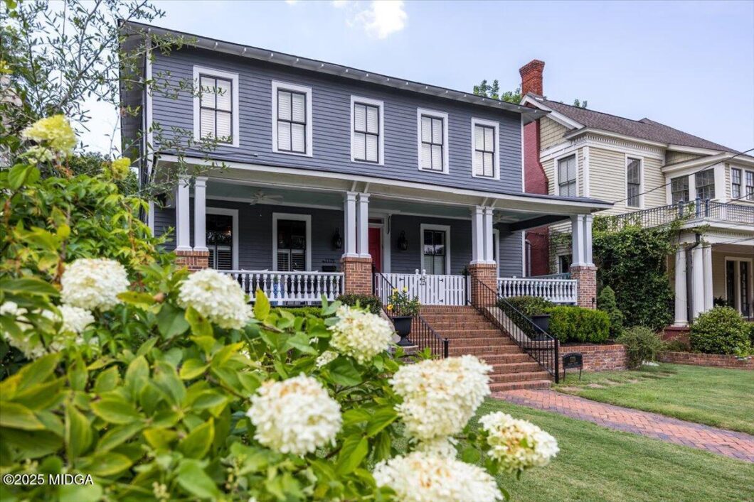 Blue two-story house with a wide covered front porch, white railings, brick steps, and blooming white hydrangeas in the foreground.