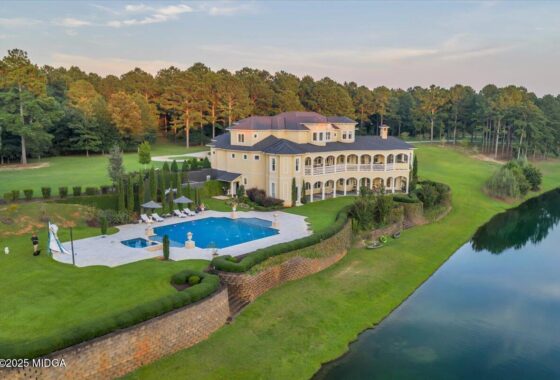 Luxurious two-story mansion by a river with a large rectangular pool and manicured grounds overlooking water.
