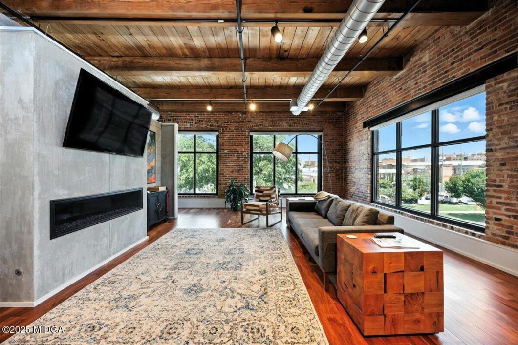 Industrial-style living room with brick walls, wooden ceiling, and large windows; gray sofa, wooden block side table, TV mounted on concrete wall, and patterned rug.