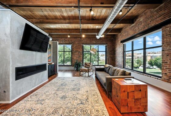 Industrial-style living room with brick walls, wooden ceiling, and large windows; gray sofa, wooden block side table, TV mounted on concrete wall, and patterned rug.