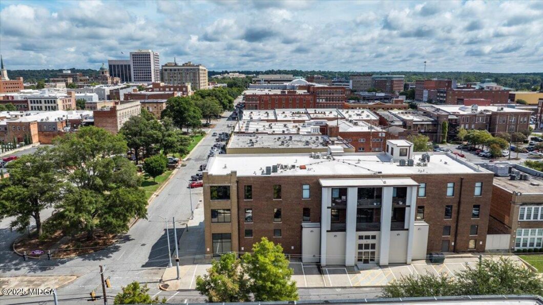 Aerial view of a brick city block with a prominent three-column white entrance, tree-lined street, and surrounding low-rise buildings under a cloudy sky.
