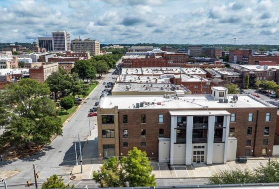 Aerial view of a brick city block with a prominent three-column white entrance, tree-lined street, and surrounding low-rise buildings under a cloudy sky.