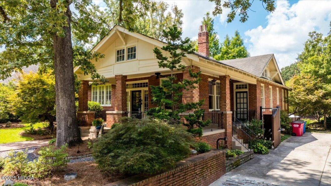 Brick bungalow with a front porch supported by brick columns, surrounded by trees on a sunny day.