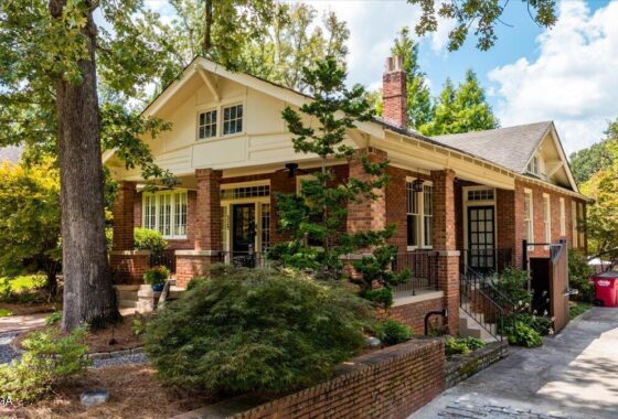 Brick bungalow with a front porch supported by brick columns, surrounded by trees on a sunny day.