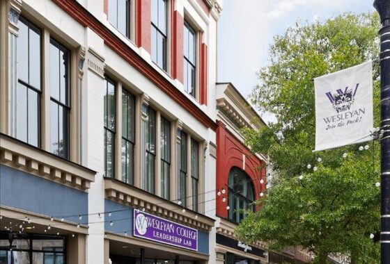 Urban street scene with brick storefronts; a Wesleyan College Leadership Lab sign is above the entrance. A white flag with the Wesleyan logo hangs by a tree on the right.