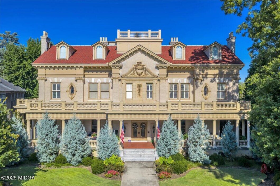 Front view of a grand beige historic mansion with a red roof, wraparound porch, and American flags in the yard.