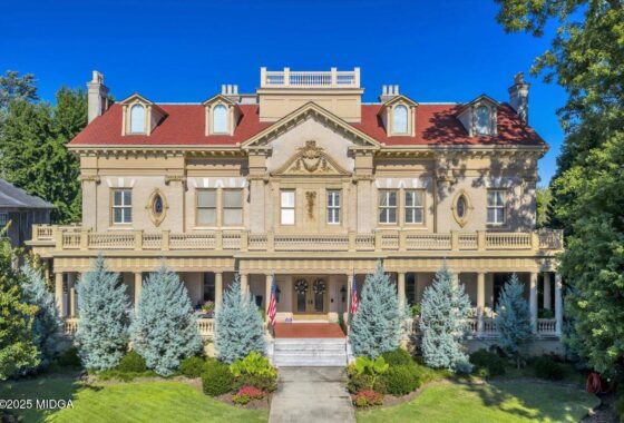 Front view of a grand beige historic mansion with a red roof, wraparound porch, and American flags in the yard.