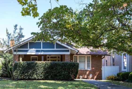 Brick bungalow with a covered front porch, blue gable trim, and a tall hedge in front. If there’s a label: avoid extra details.