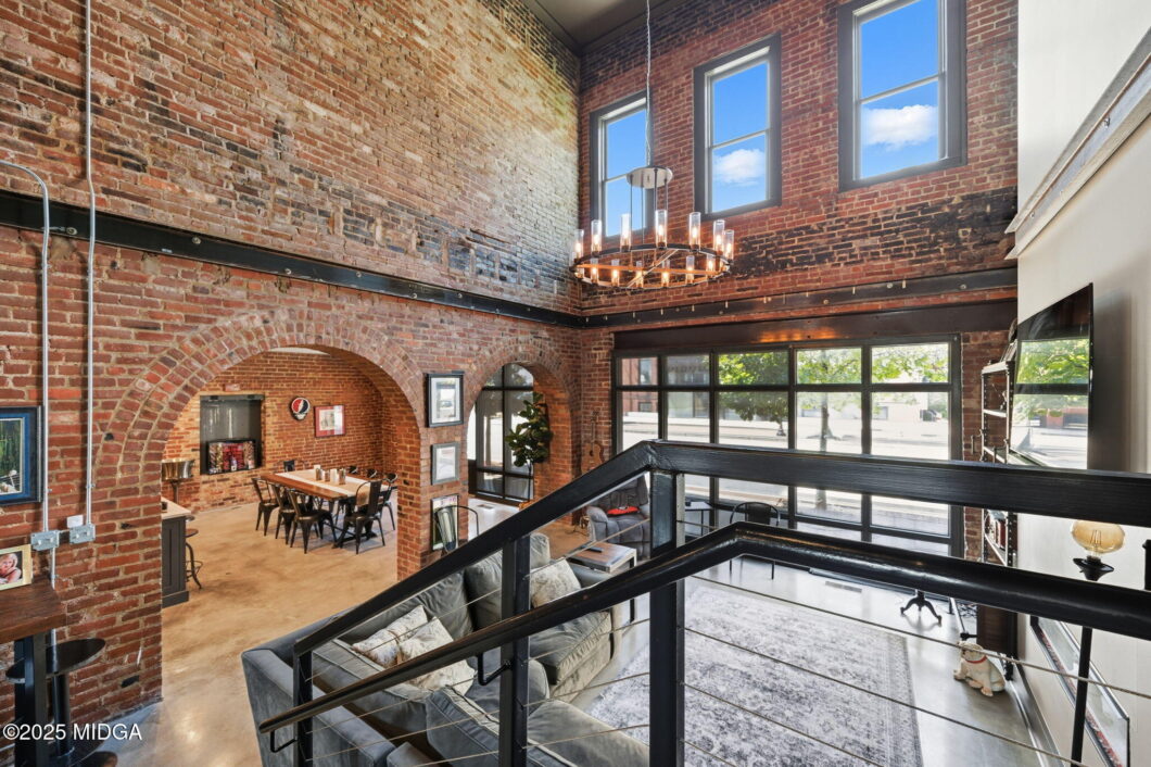 Industrial loft interior with exposed brick walls, arched brick doorways, and a circular chandelier seen from a mezzanine level.